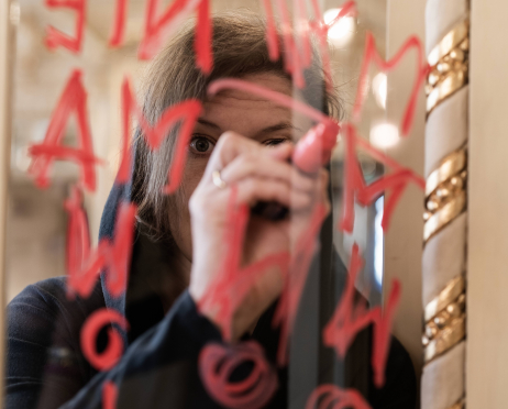 Martyna Krzysztofik stoi przy lustrze w ozdobnym wnętrzu teatru. Ma na sobie czarną bluzę z kapturem założonym na głowę i trzyma w dłoni czerwony marker, którym pisze na szybie duże, odręczne litery. Twarz częściowo zasłania ręka i napis. W tle widoczne są złote zdobienia i rozmyte światła wnętrz.